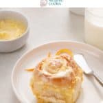 Frosted orange roll on a white plate with a glass of milk and the pan of rolls in the background. Text overlay includes recipe name.