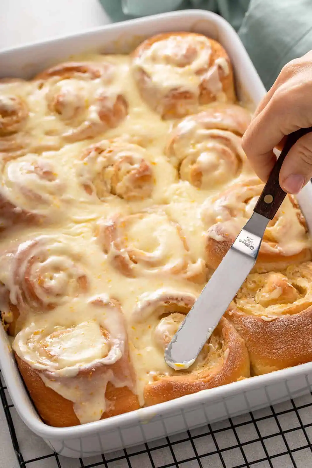 Offset spatula spreading orange icing over the top of baked orange rolls.