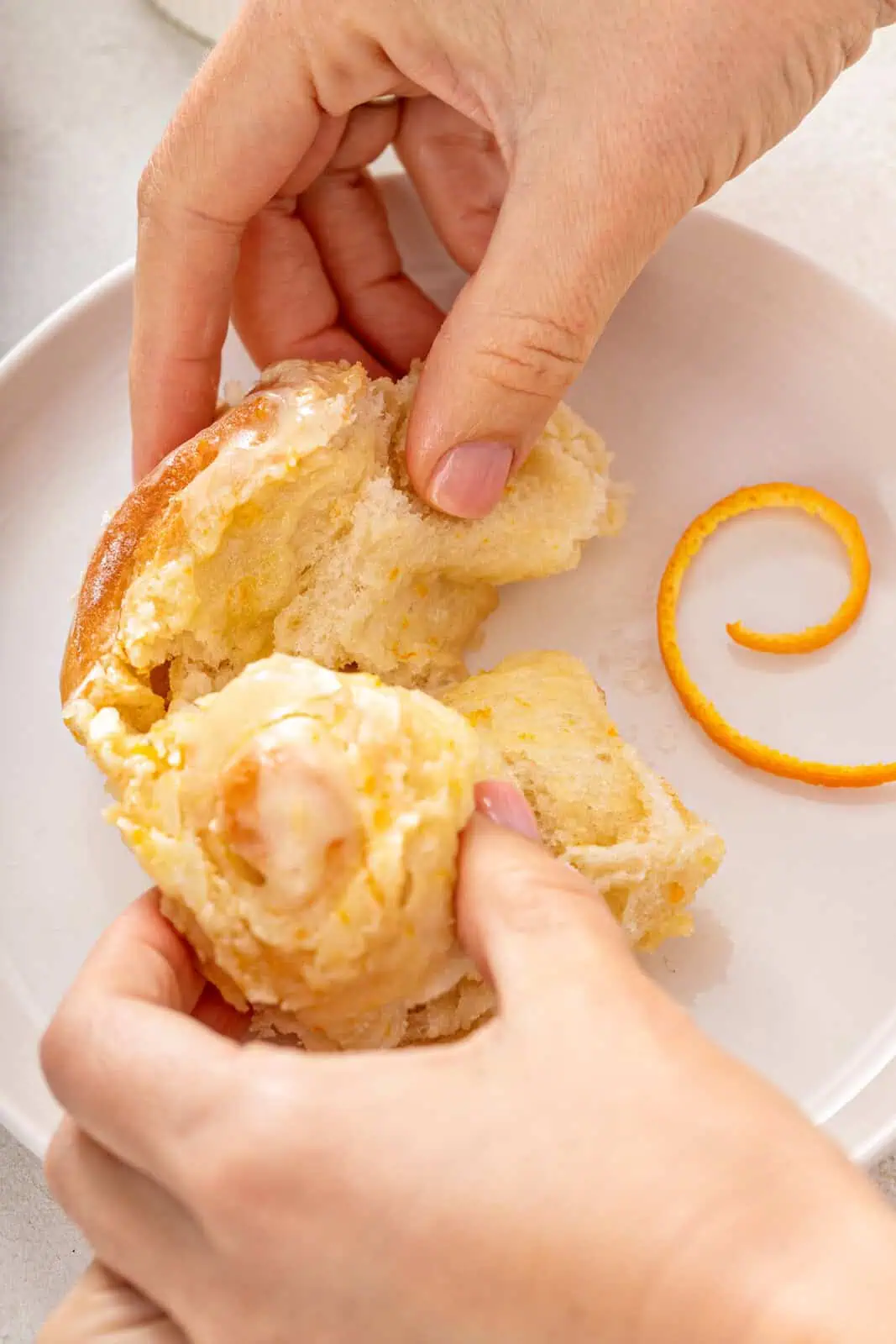 Hand pulling apart a frosted orange roll on a white plate.