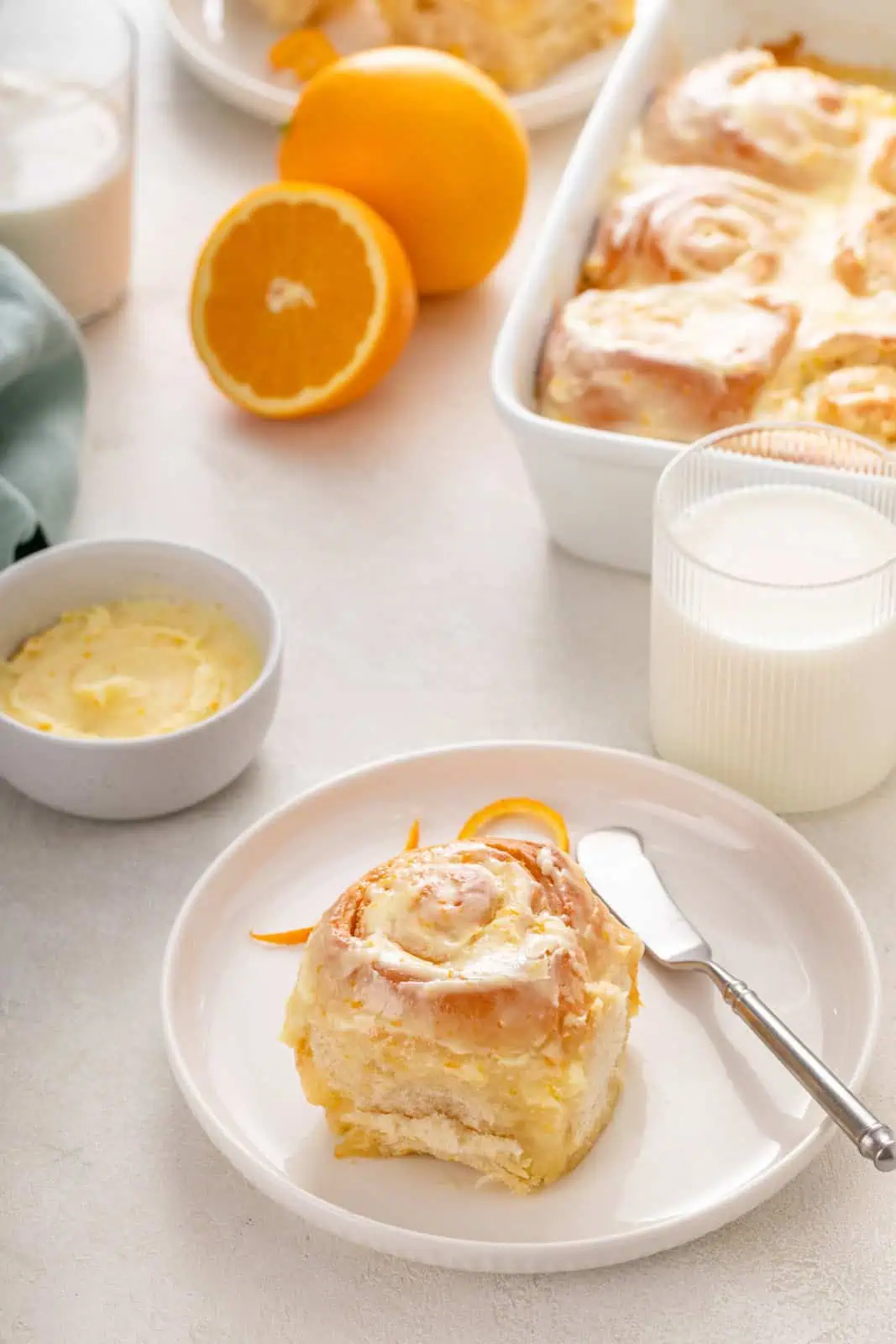 Frosted orange roll on a white plate with a glass of milk and the pan of rolls in the background.