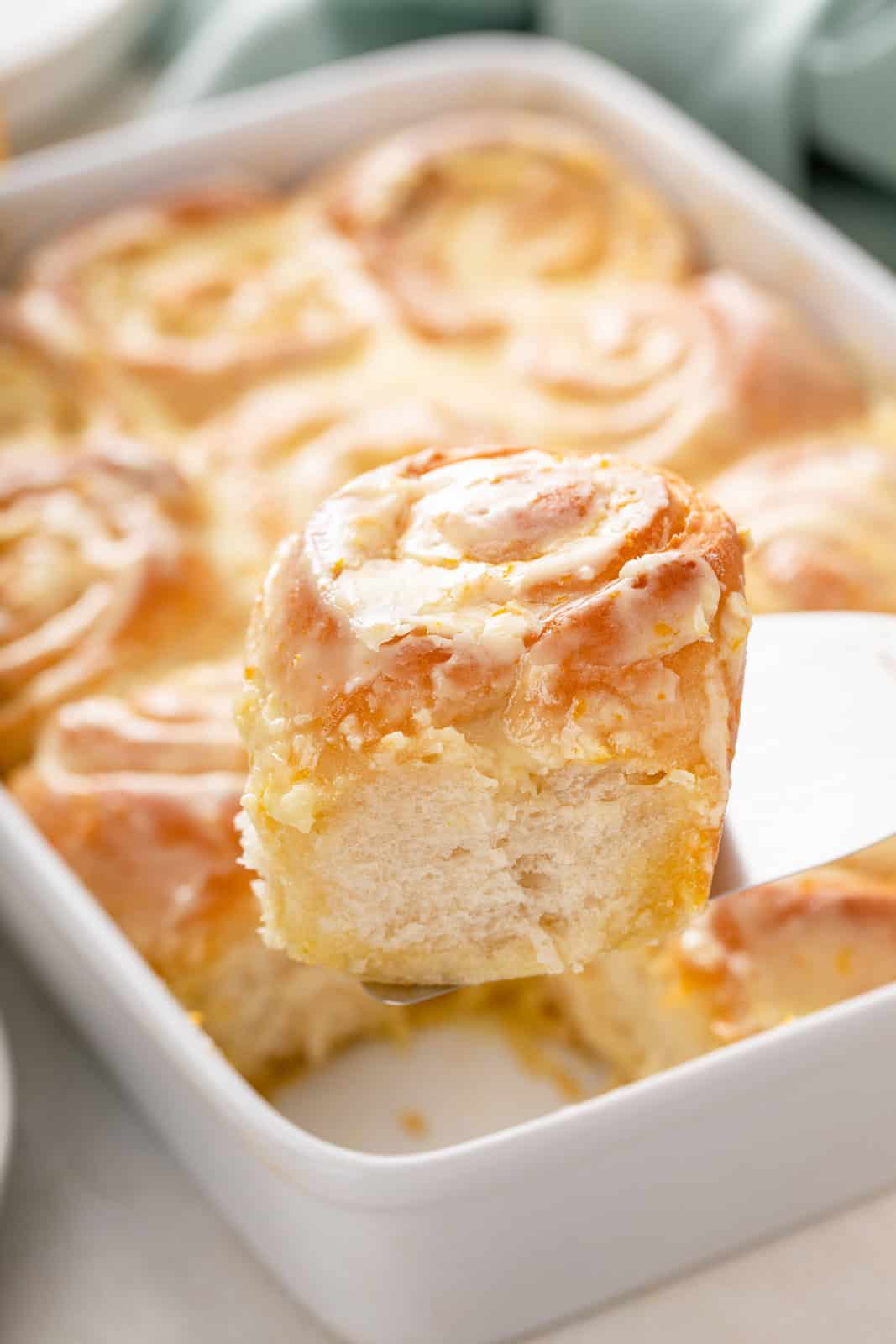 Cake server lifting up a frosted orange roll from the baking dish.