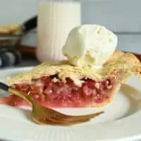 Slice of rhubarb pie topped with a scoop of vanilla ice cream, set next to a gold fork on a white plate.