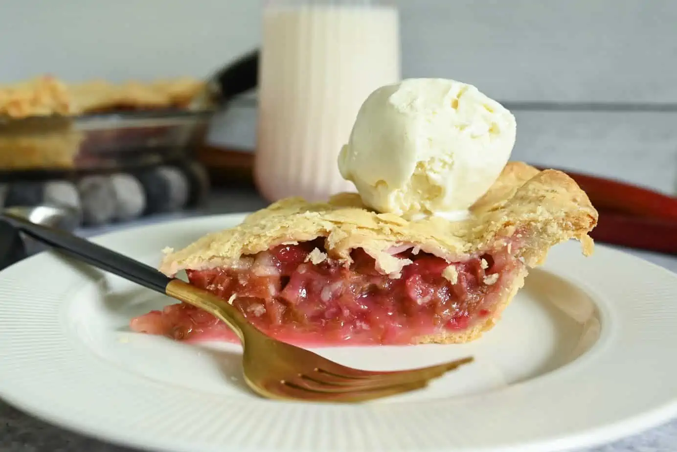 Slice of rhubarb pie on a white plate. The pie is topped with ice cream and is set next to a gold fork.