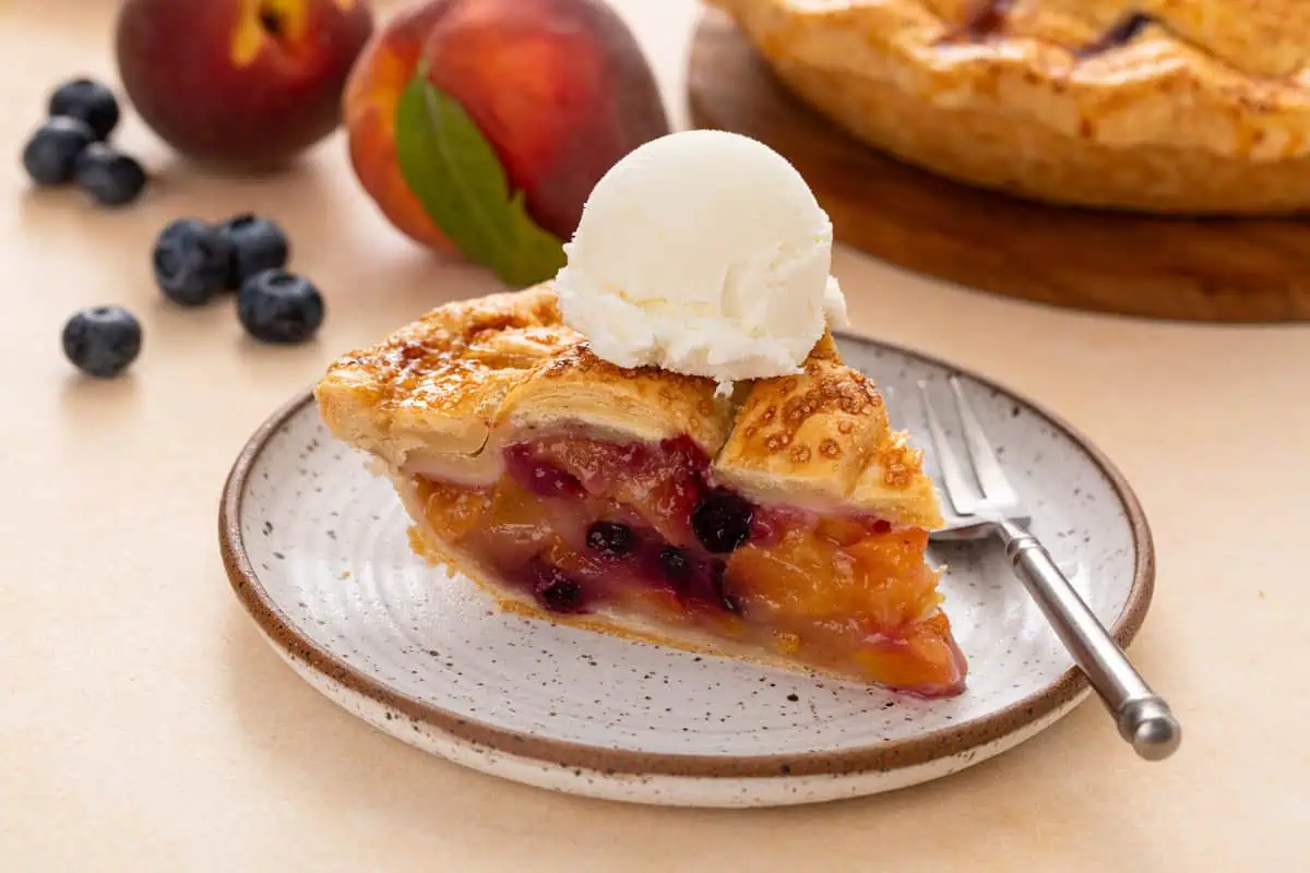 Plated slice of peach blueberry pie topped with a scoop of vanilla ice cream.