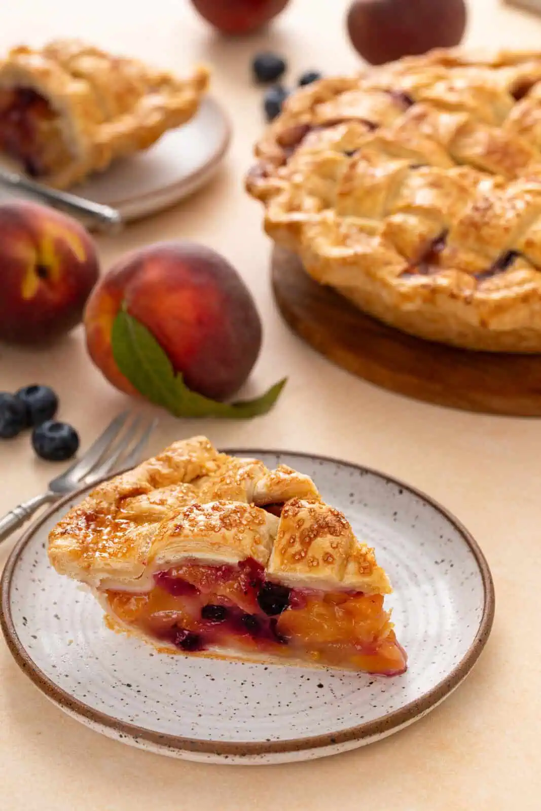 Slice of peach blueberry pie on a plate with the rest of the pie in the background.