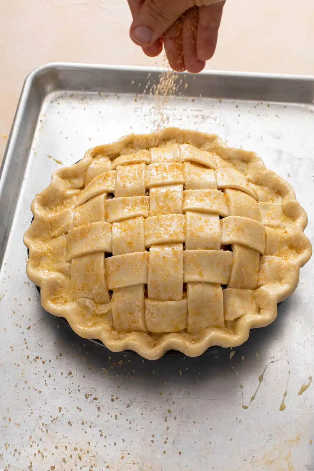 Lattice-topped pie on a baking sheet with a hand sprinkling coarse sugar over the top.