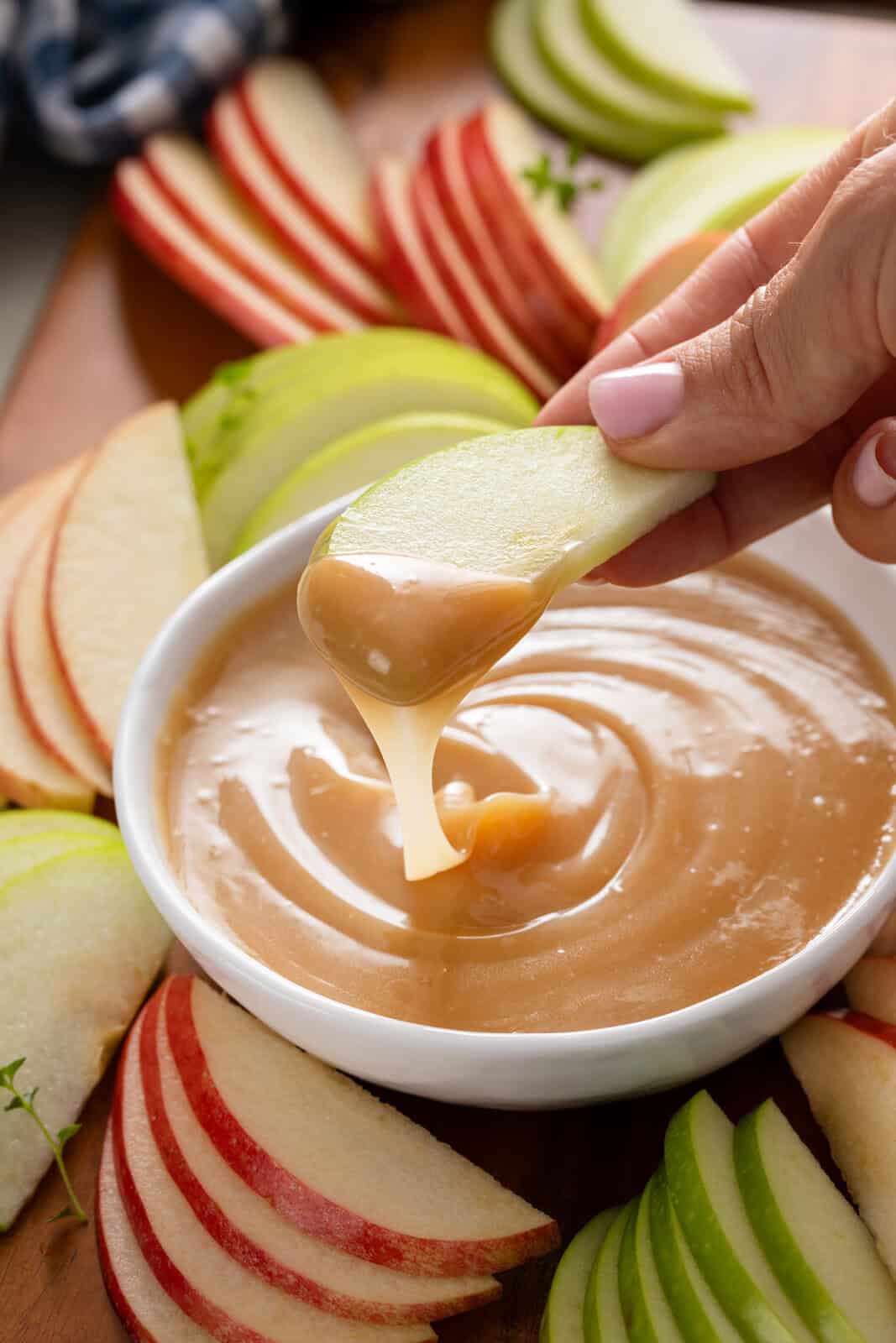 Apple slice dipping in caramel apple dip being held over a bowl of the dip.