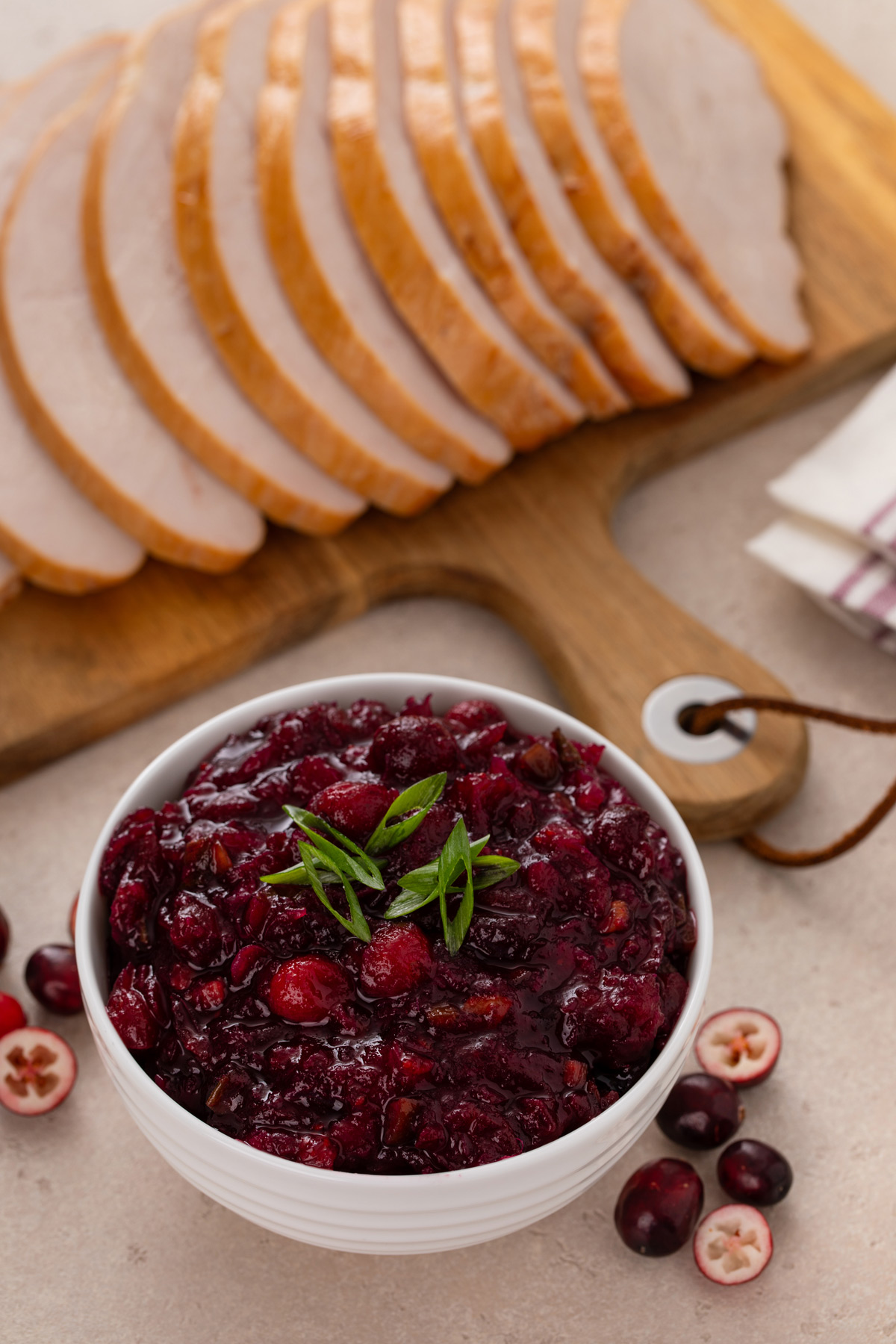 White bowl of cranberry chutney on a countertop next to a board of sliced turkey breast.