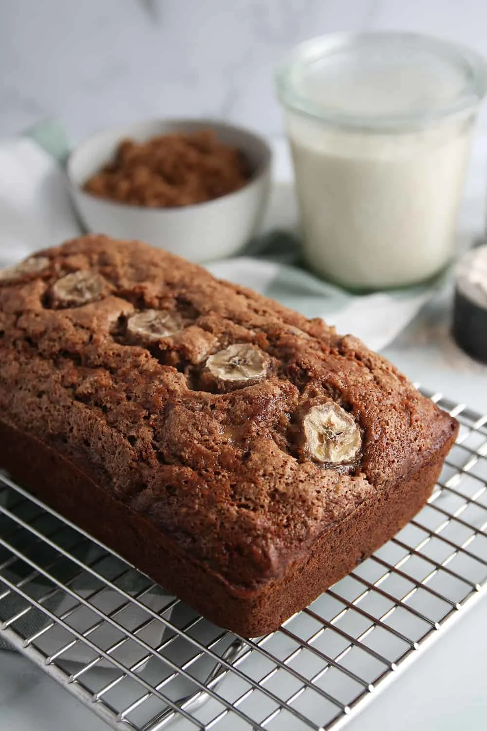 Baked loaf of vegan sourdough banana bread on a cooling rack, with a bowl of brown sugar and a glass jar of sourdough starter in the background