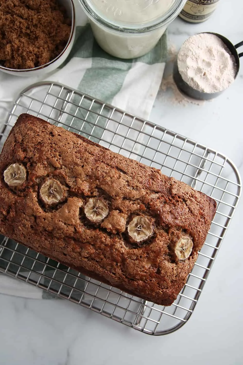 Overhead view of baked loaf of vegan sourdough banana bread on a cooling rack next to a bowl of brown sugar, a measuring cup of flour and a jar of sourdough starter