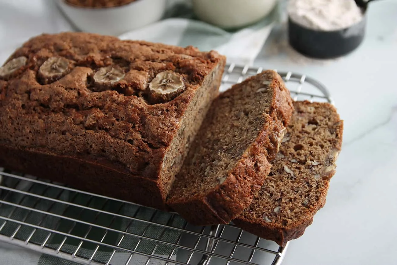 Angled view of sliced loaf of sourdough banana bread on a cooling rack with a measuring cup of flour in the backgroun