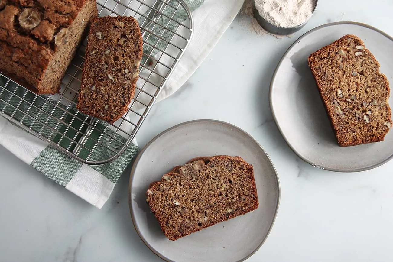 Overhead view of 2 gray plates with slices of sourdough banana bread next to a sliced loaf of banana bread on a cooling rack