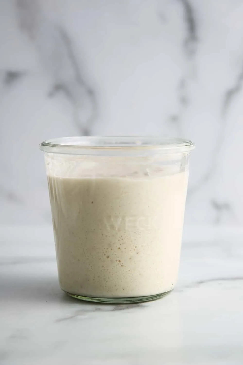 Side view of bubbly sourdough starter in a glass jar in front of a marble backsplash