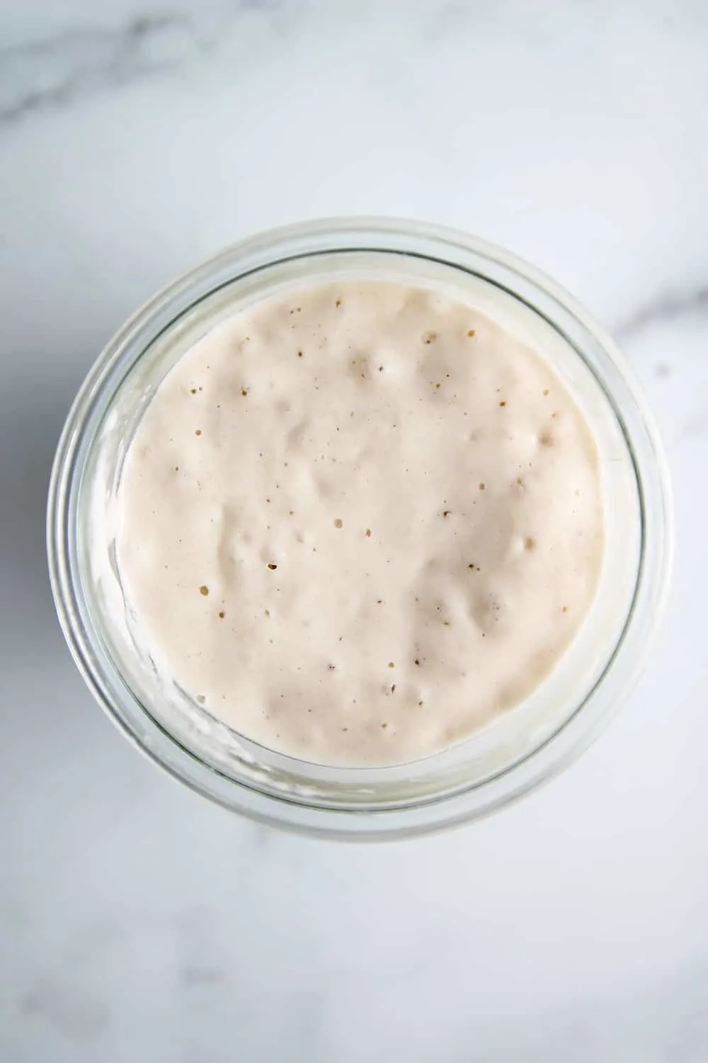 Overhead view of bubbly sourdough starter in a glass jar on a marble counter