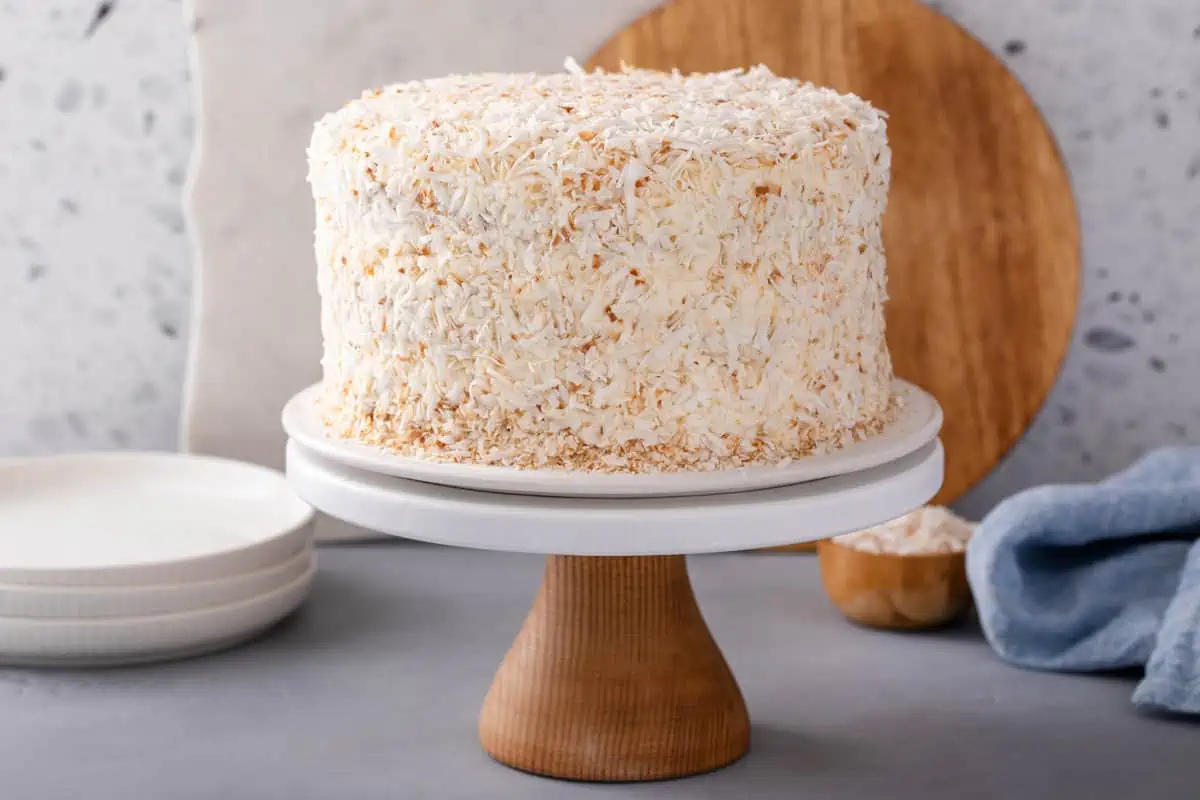 Coconut cake covered in toasted coconut on a white and wood cake stand.