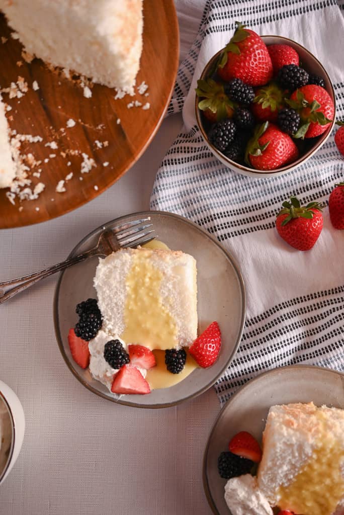 Slice of angel food cake on a gray plate, topped with lemon curd, berries, and whipped cream