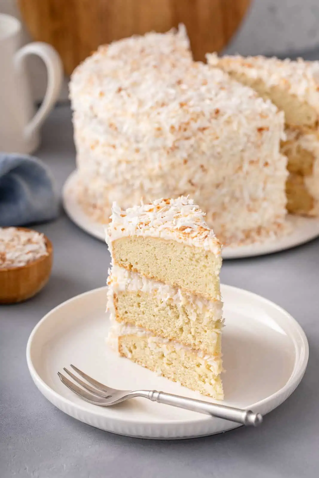 Slice of coconut cake on a white plate with the rest of the cake in the background.
