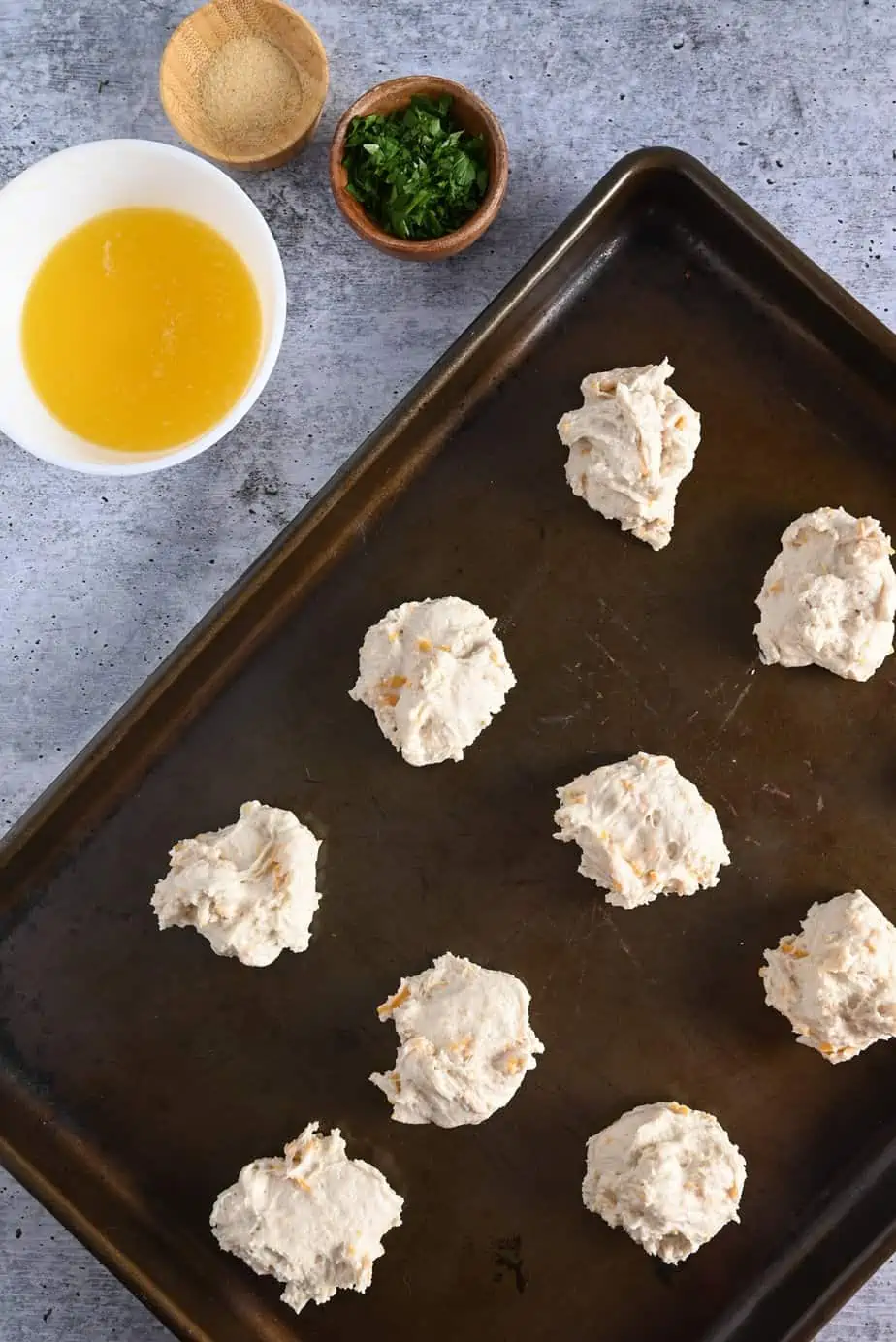 Unbaked cheddar bay biscuits on a baking sheet next to bowls of melted butter, garlic powder, and minced parsley.