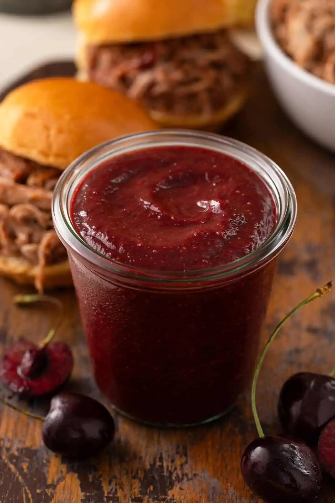 Close up of cherry barbecue sauce on a wooden board with pulled pork sandwiches in the background.