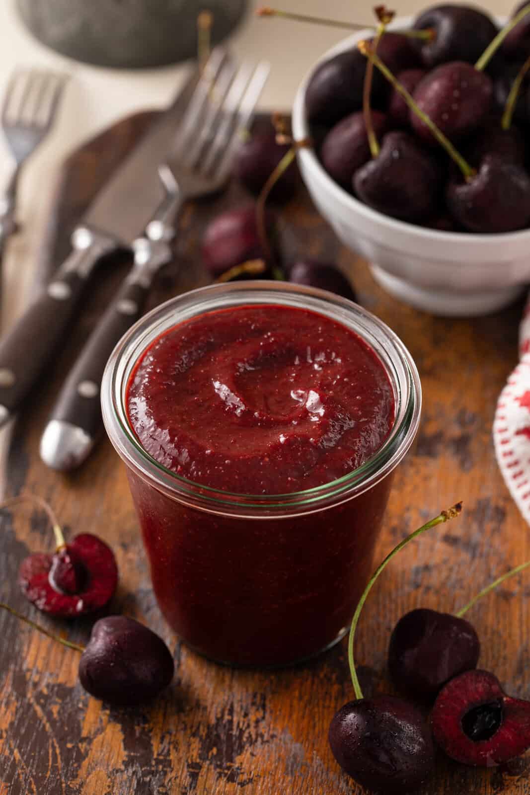 Glass jar filled with cherry barbecue sauce with a bowl of sweet cherries in the background.