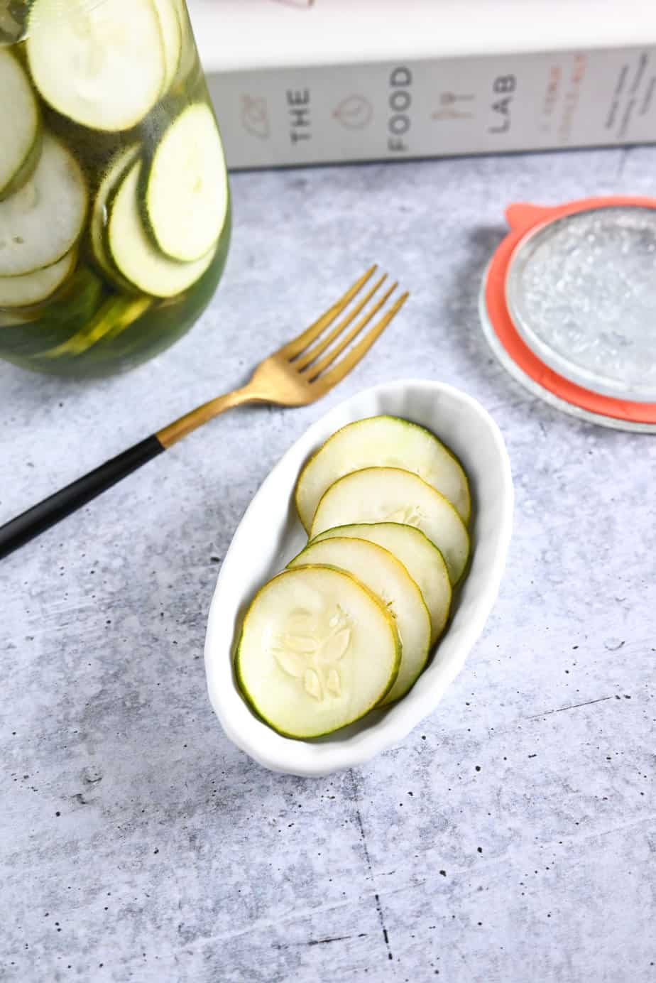 White dish with sliced pickled cucumbers, set next to a gold fork on a concrete countertop.