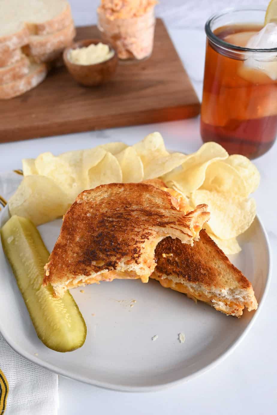 Bite taken from one half of a grilled pimento cheese sandwich on a white plate, next to a pickle and potato chips.