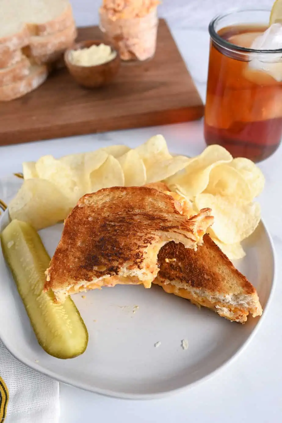 Bite taken from one half of a grilled pimento cheese sandwich on a white plate, next to a pickle and potato chips.