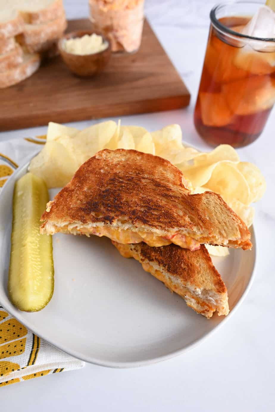 Two halves of a grilled pimento cheese sandwich arranged on a white plate with potato chips and a pickle spear.