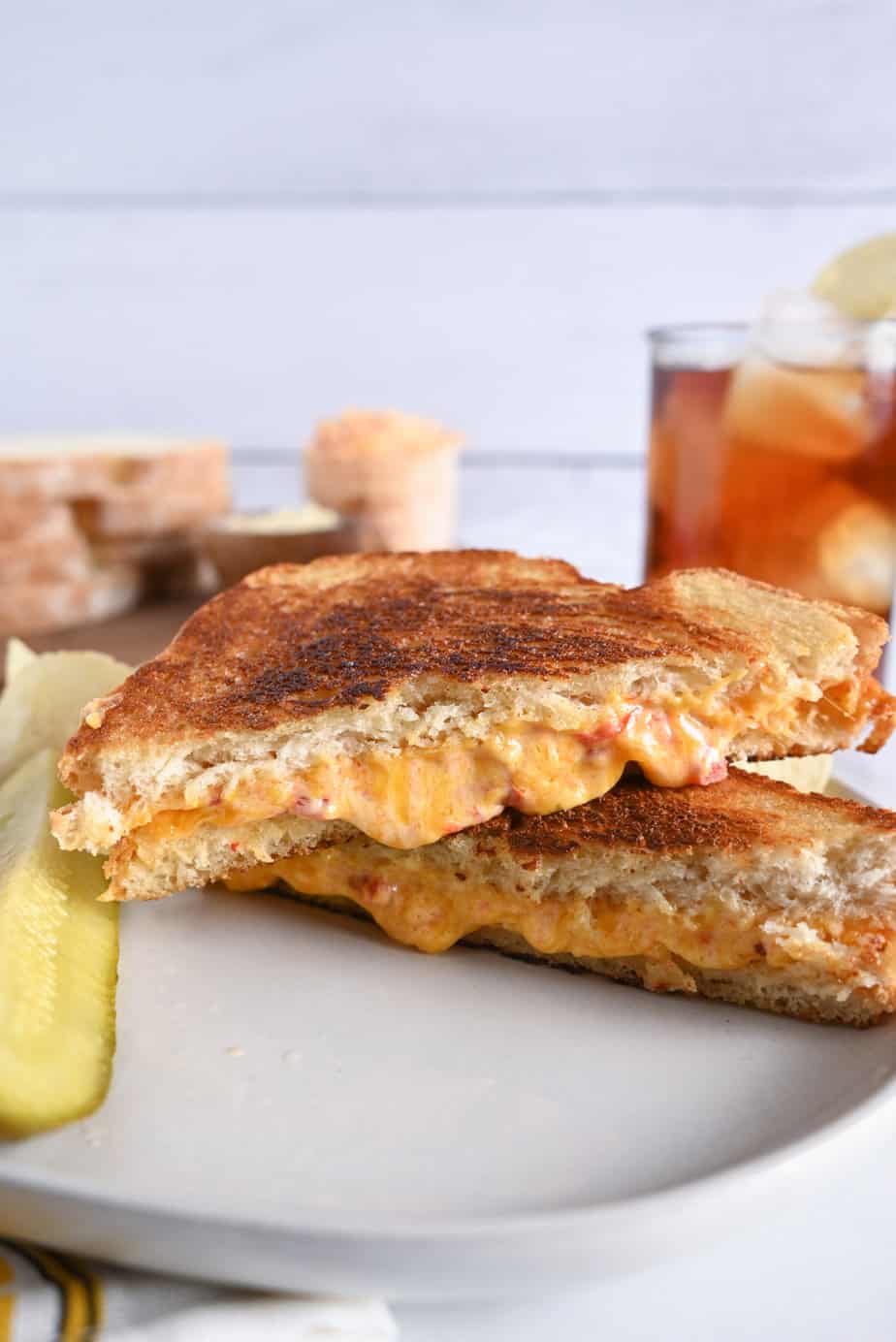 Side view of two halves of a grilled pimento cheese sandwich stacked on a white plate. A glass of iced tea is visible in the background.