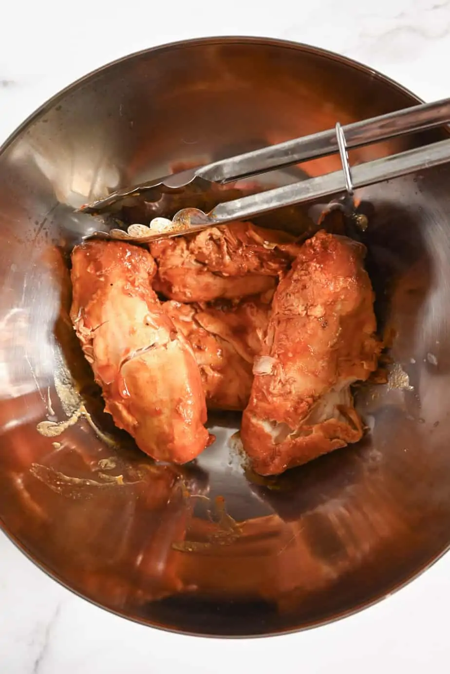 Slow cooked barbecue chicken breasts in a metal bowl with metal tongs.