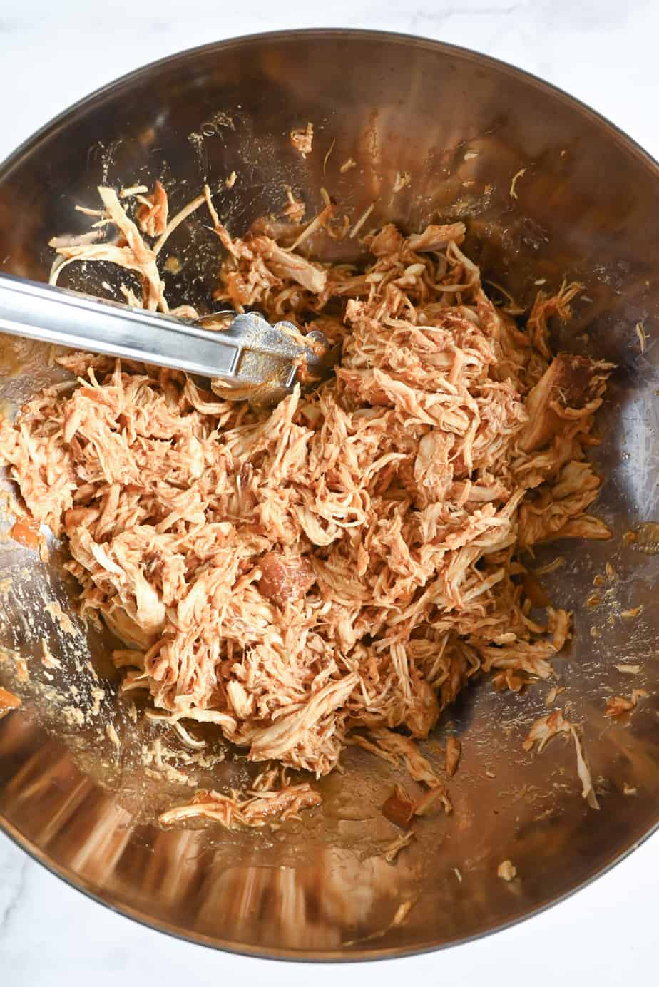 Shredded chicken mixed with barbecue sauce in a metal mixing bowl.