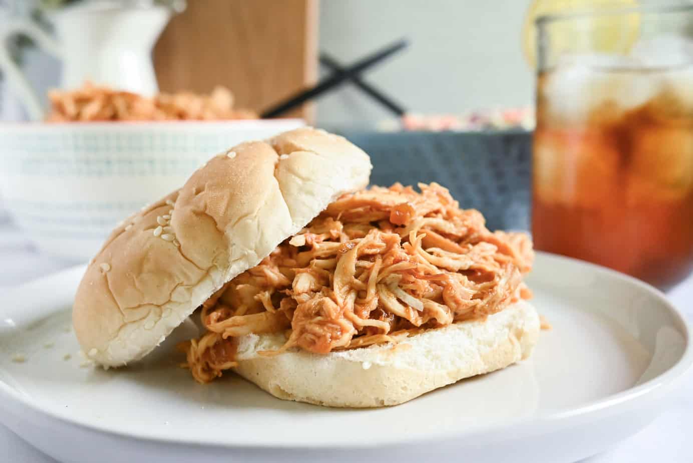 Shredded barbecue chicken on a bun, with iced tea and bowls of chicken and slaw in the background.