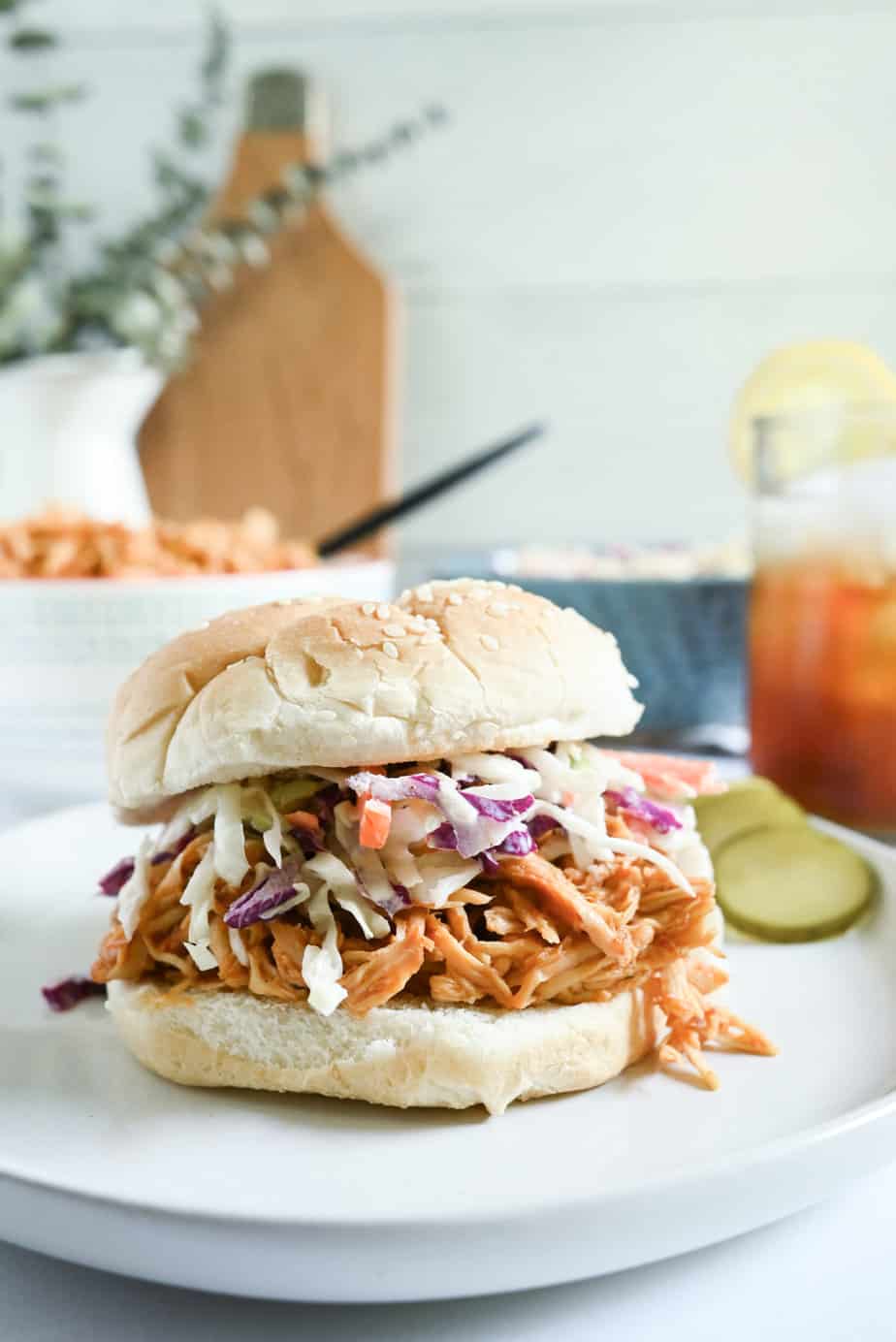 Crockpot pulled chicken sandwich on a bun, topped with slaw, next to a glass of iced tea.