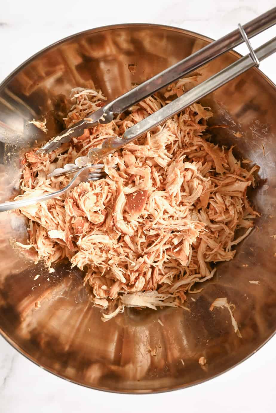 Shredded slow cooked chicken in a metal bowl with a pair of tongs.