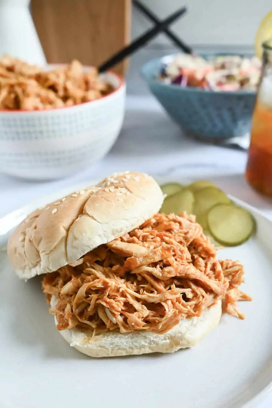 Crockpot pulled chicken on a sesame bun, set next to pickles on a white plate.