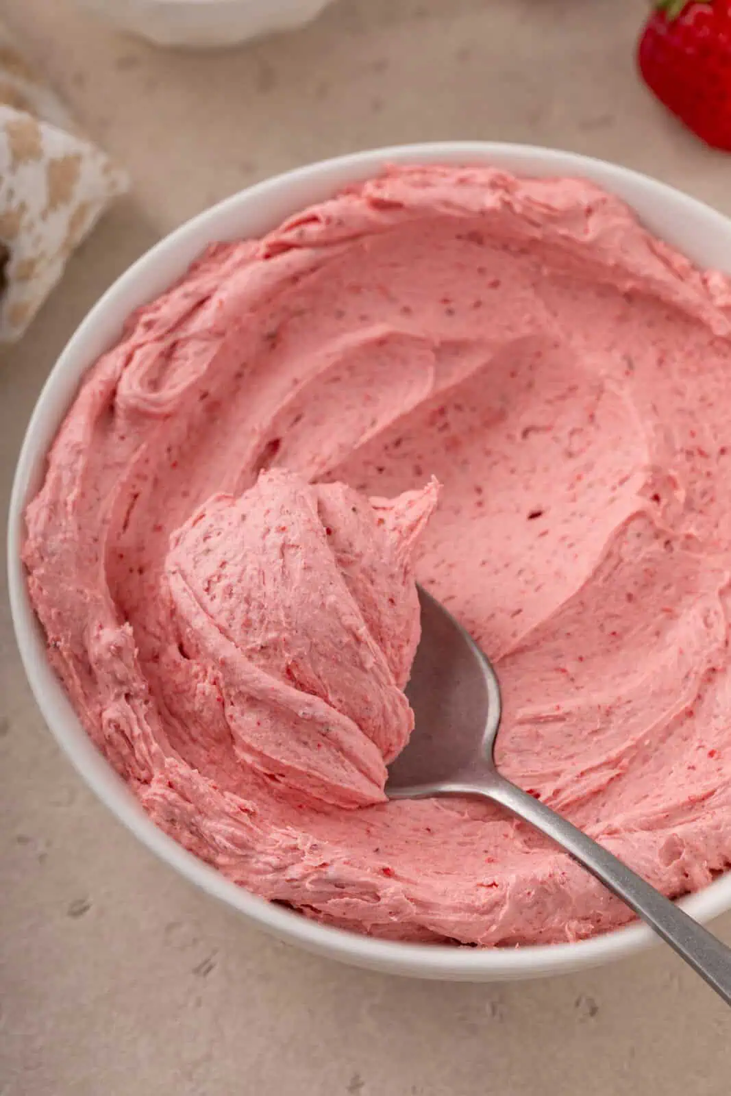 Spoon lifting up a bite of strawberry buttercream frosting from a white bowl.