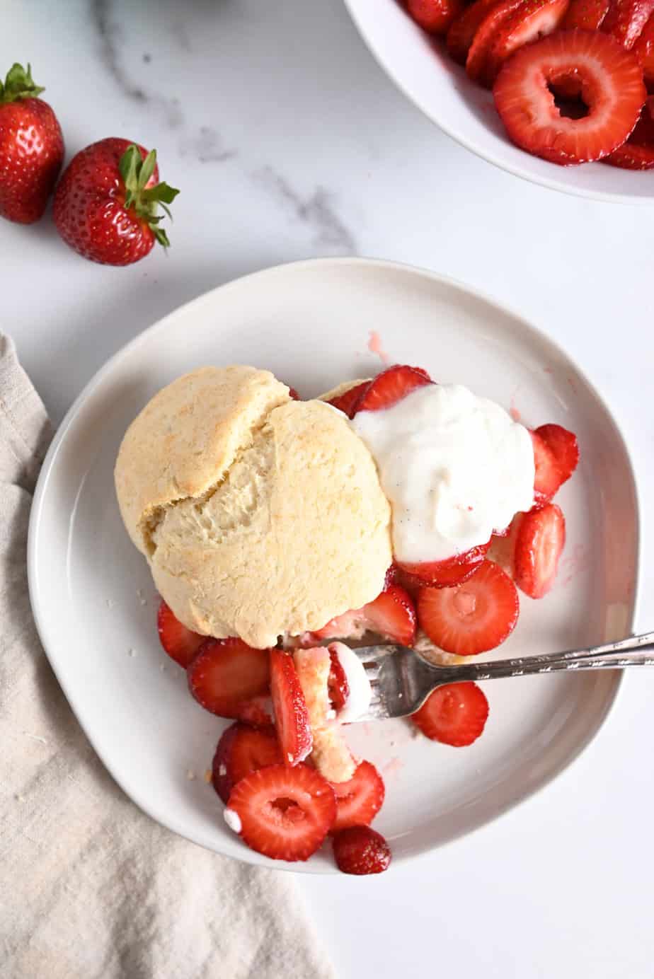 Overhead view of strawberry shortcake on a white plate, with a fork taking a bite of the cake, strawberries, and whipped cream.