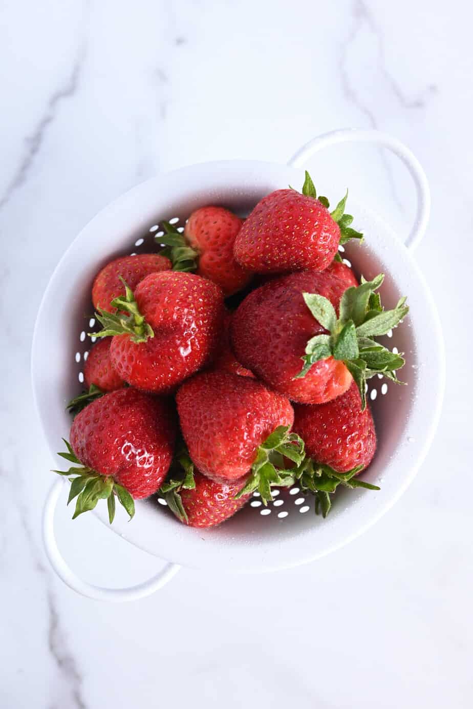 Fresh strawberries in a white colander, set on a marble countertop.