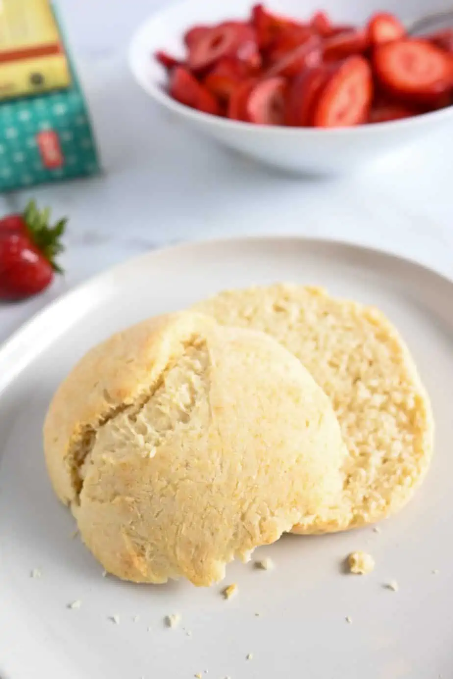 Halved shortcake biscuit set on a white plate with a bowl of strawberries in the background.