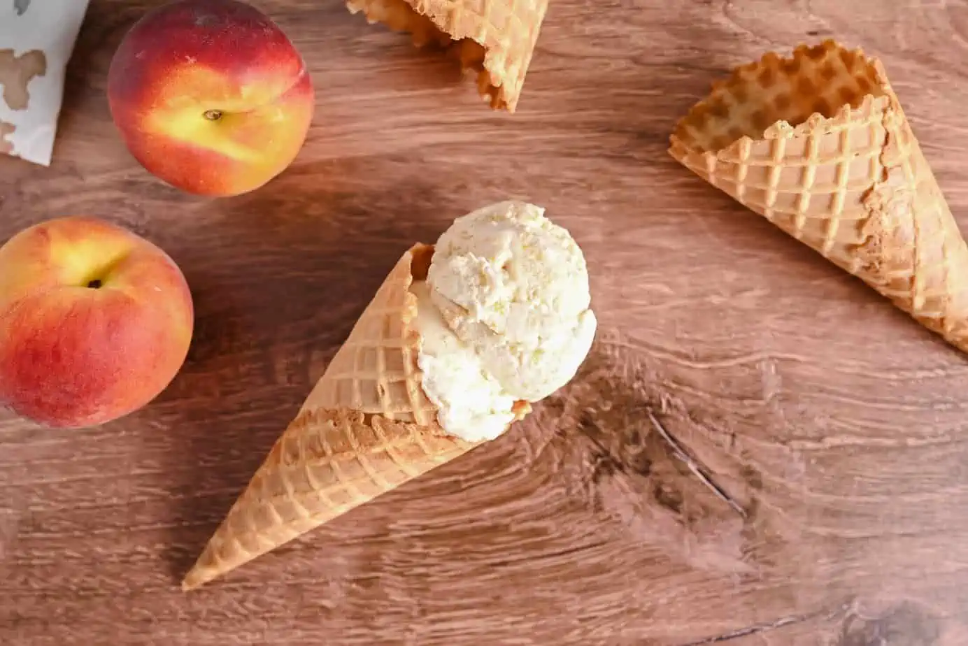 Waffle cone holding peach ice cream laying on a wooden countertop next to peaches and empty waffle cones.