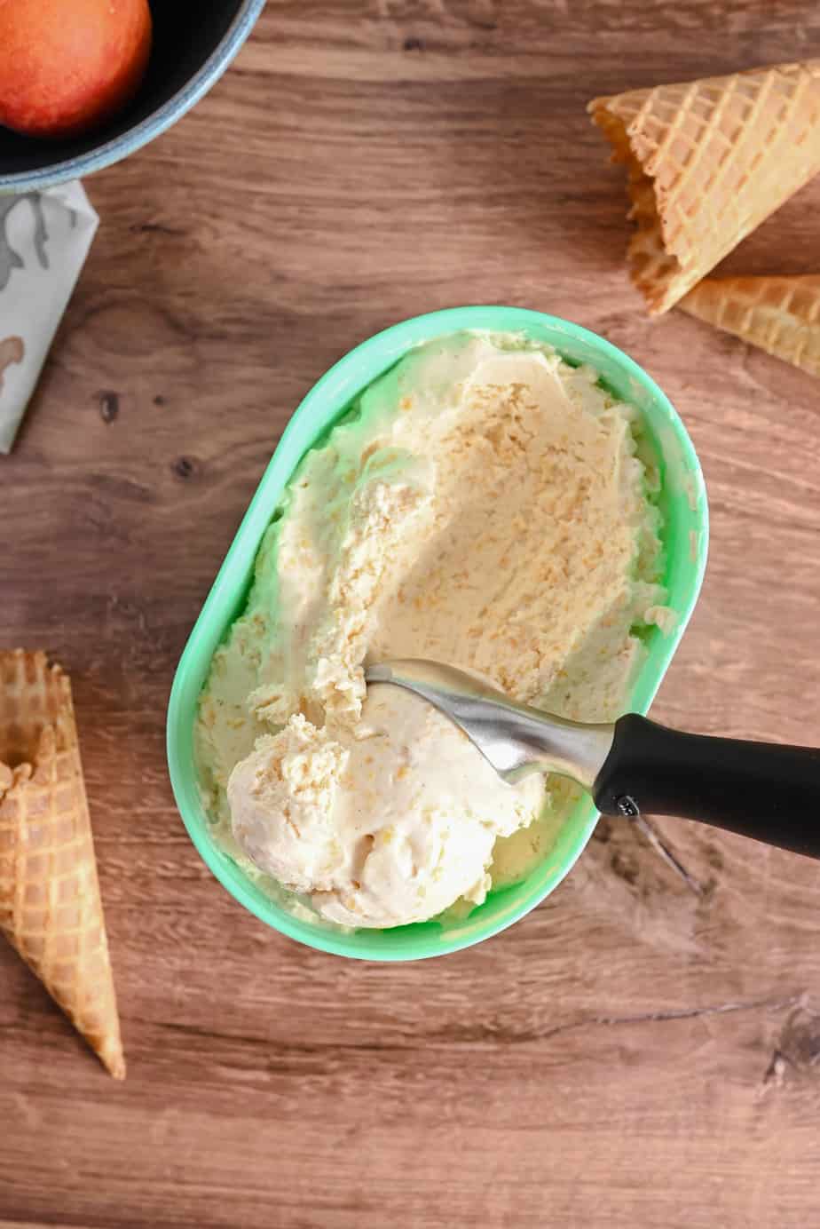 Ice cream scoop scooping peach ice cream out of a mint-colored container set on a wooden countertop.