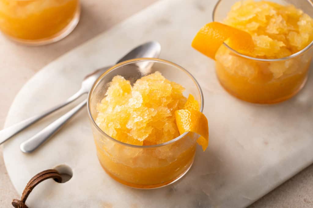 Two glasses of bourbon slush garnished with orange twists on a marble board.
