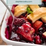 Spoon lifting a bite of mixed berry cobbler out of a baking dish. Text overlay includes recipe name.