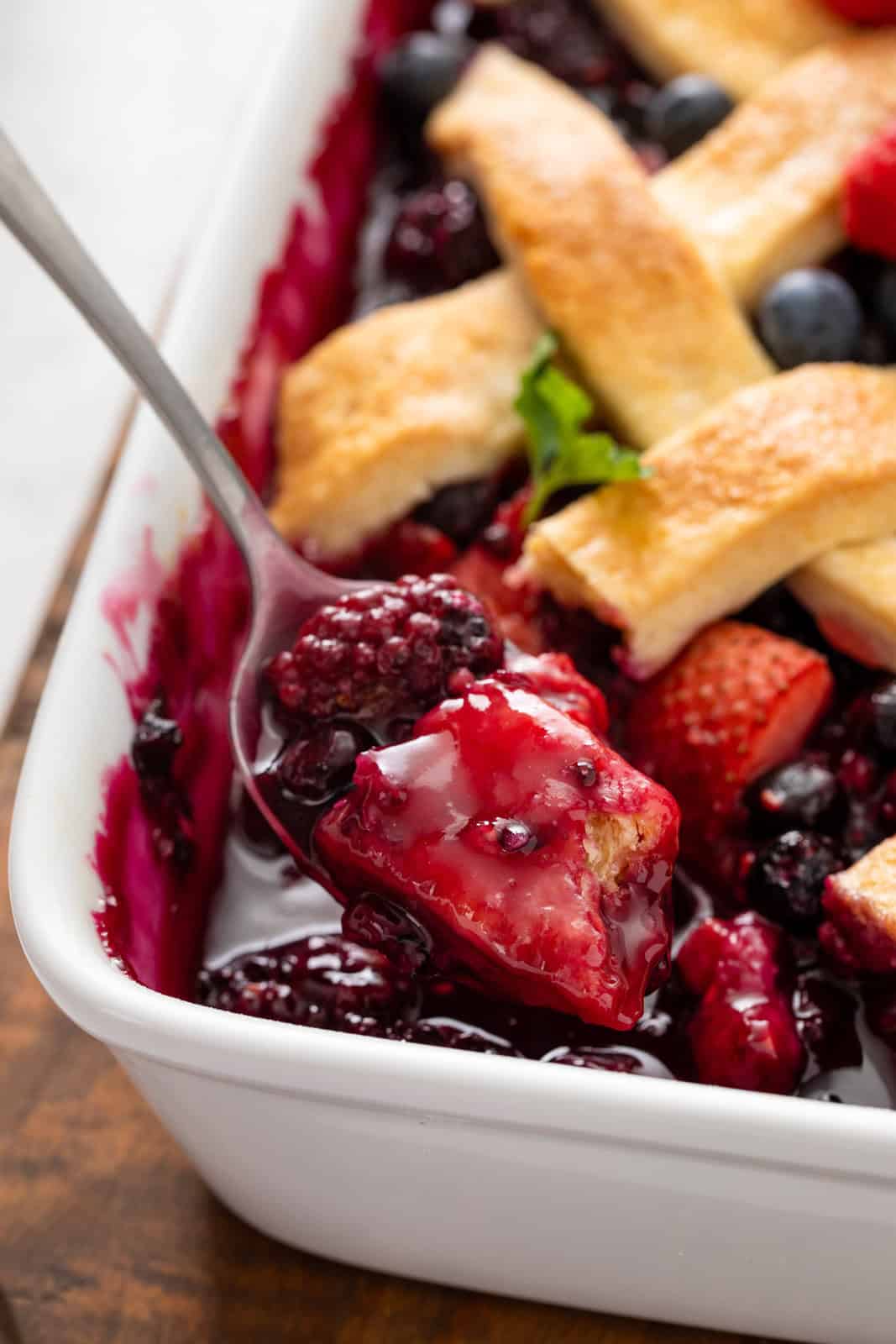 Spoon lifting a bite of mixed berry cobbler out of a baking dish.