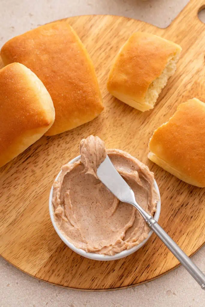 Dinner rolls surrounding a bowl of cinnamon honey butter and butter knife on a wooden board.