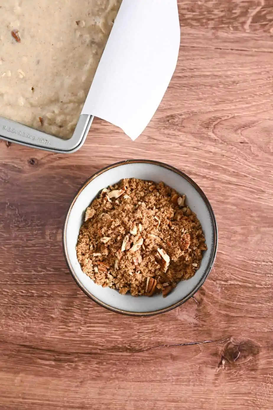 Topping for applesauce bread in a small bowl, next to a loaf pan with the bread batter in it.