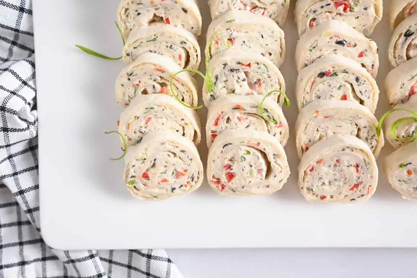 Cream cheese roll ups arranged in rows on a white platter on a black checked tea towel.