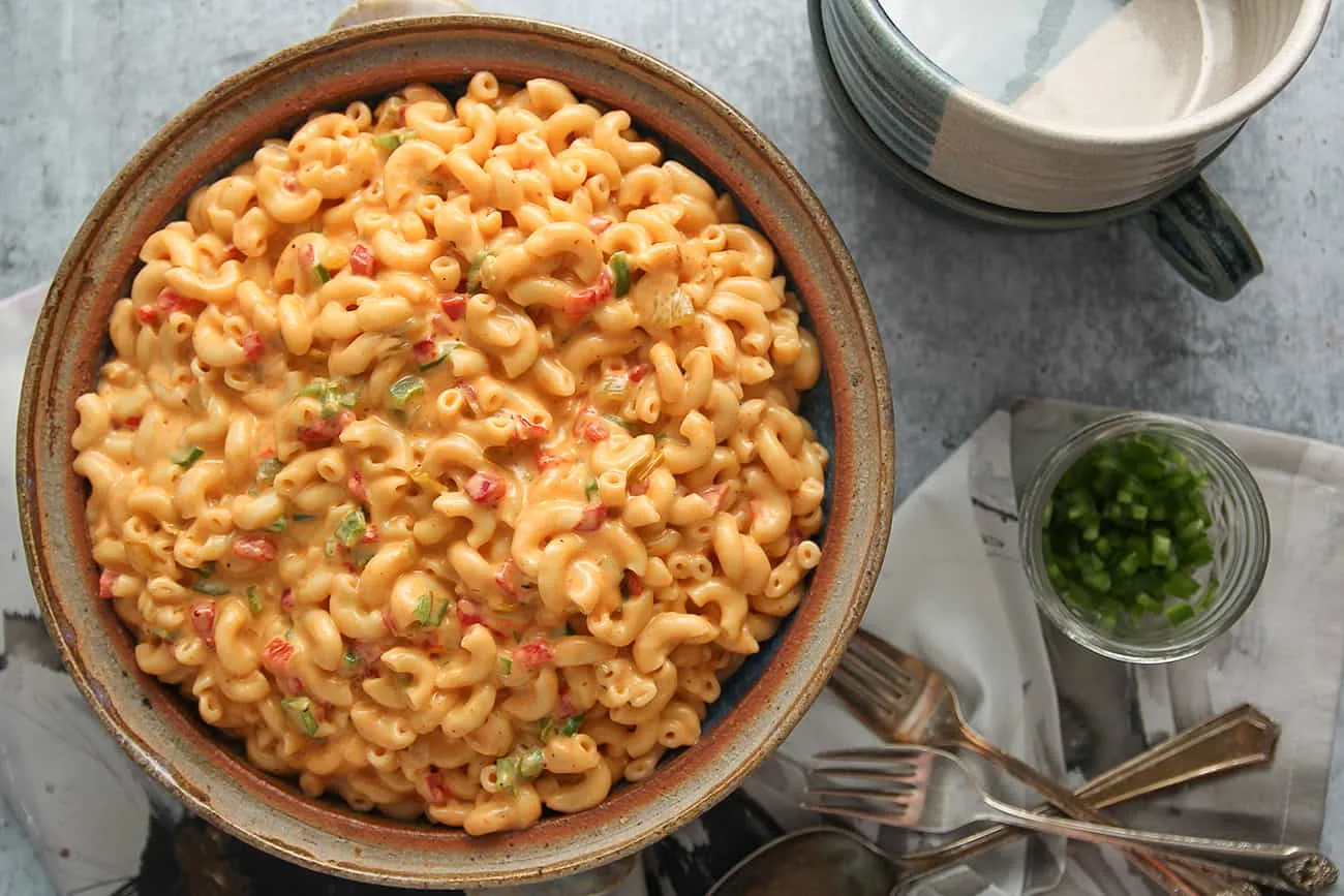 Large bowl of green chili macaroni and pimento cheese alongside serving bowls and diced jalapenos