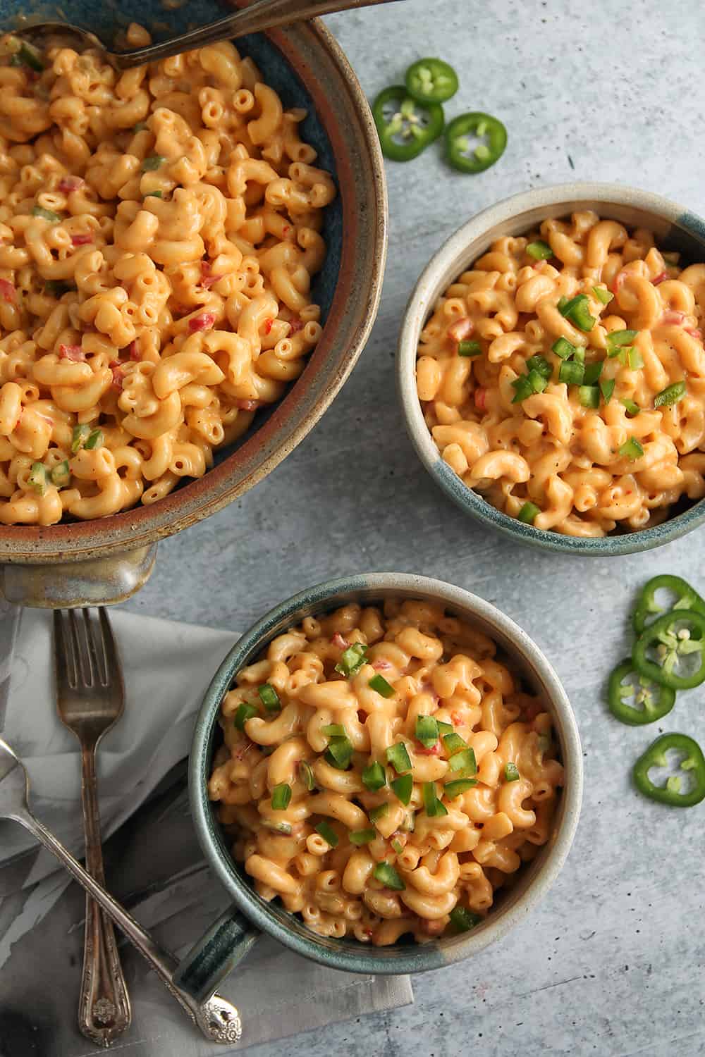 Large bowl of macaroni and pimento cheese alongside two serving bowls, forks and sliced jalapenos