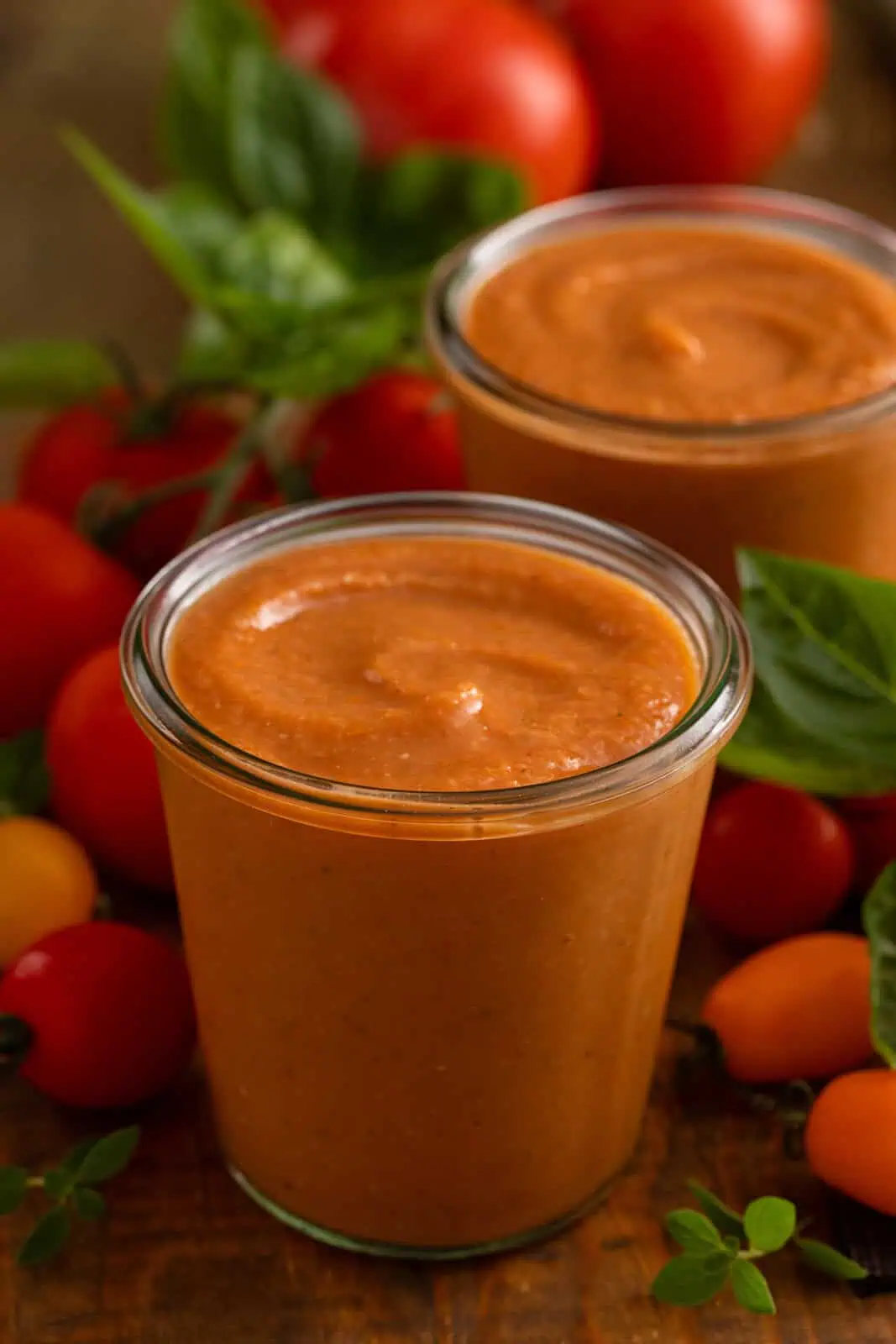 Close up of a jar filled with roasted tomato sauce, with fresh tomatoes and herbs in the background.